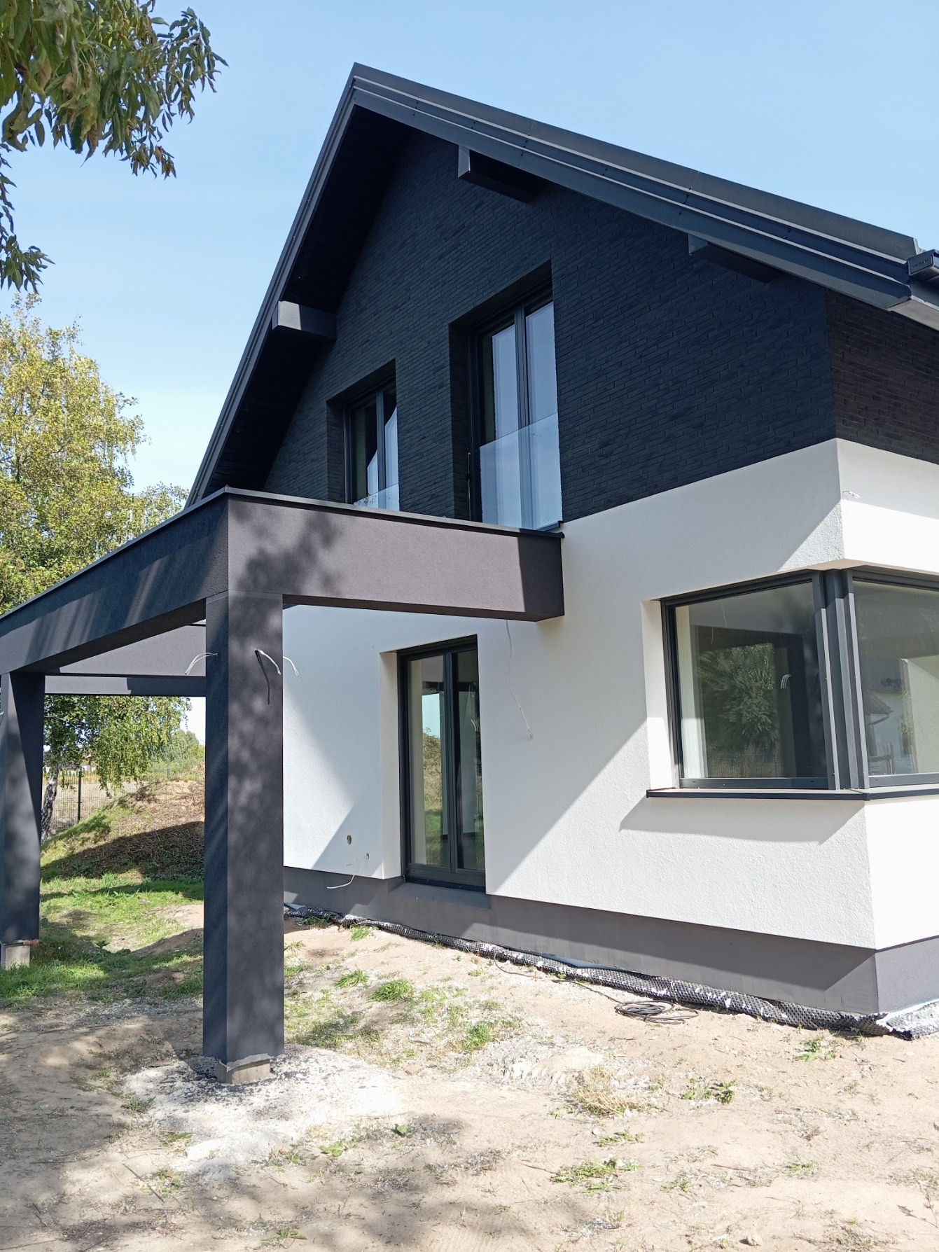 Nowoczesny dom z elewacją w dwóch odcieniach: biały tynk i grafitowa cegła. Widoczna pergola w kolorze grafitowym. Minimalistyczny design, jasne, naturalne światło.