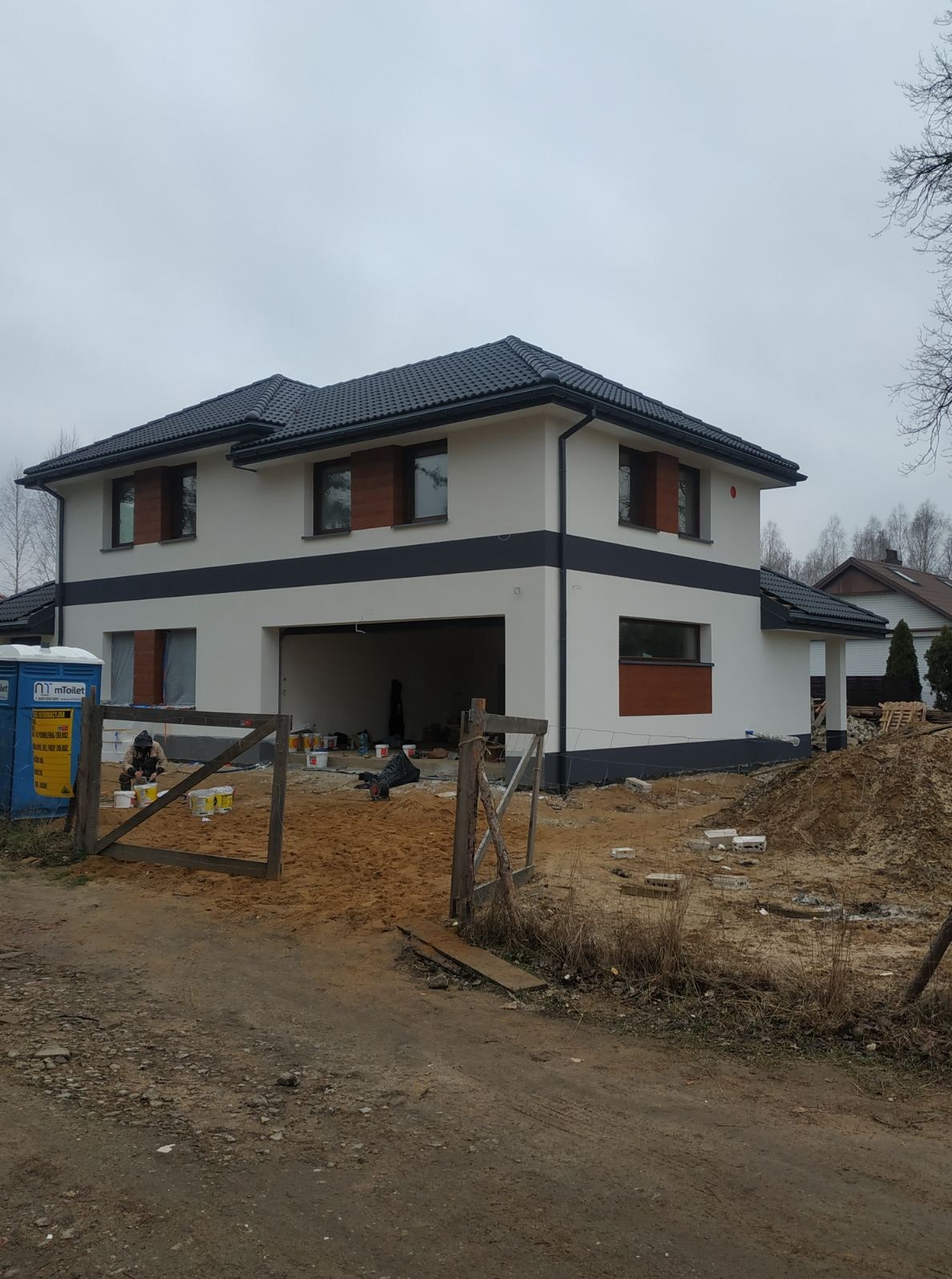 Dwukondygnacyjny dom z garażem po malowaniu elewacji na biało z ciemnymi pasami. Widoczne otwarte puszki z farbą i pracownicy na terenie budowy.