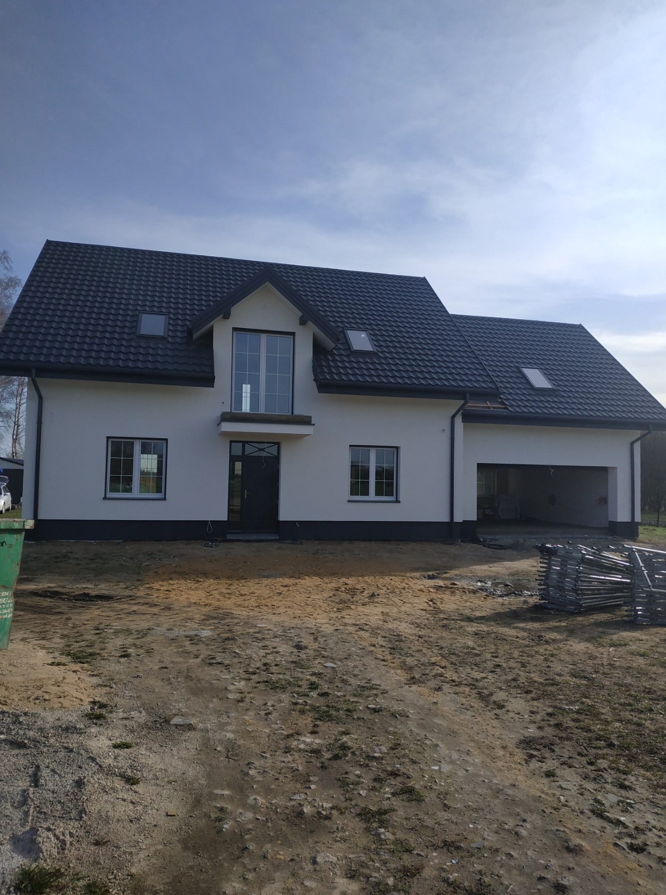 Biały dom jednorodzinny z ciemną dachówką i szarą podmurówką w słoneczny dzień. Widoczne rusztowania. Elewacja po renowacji.