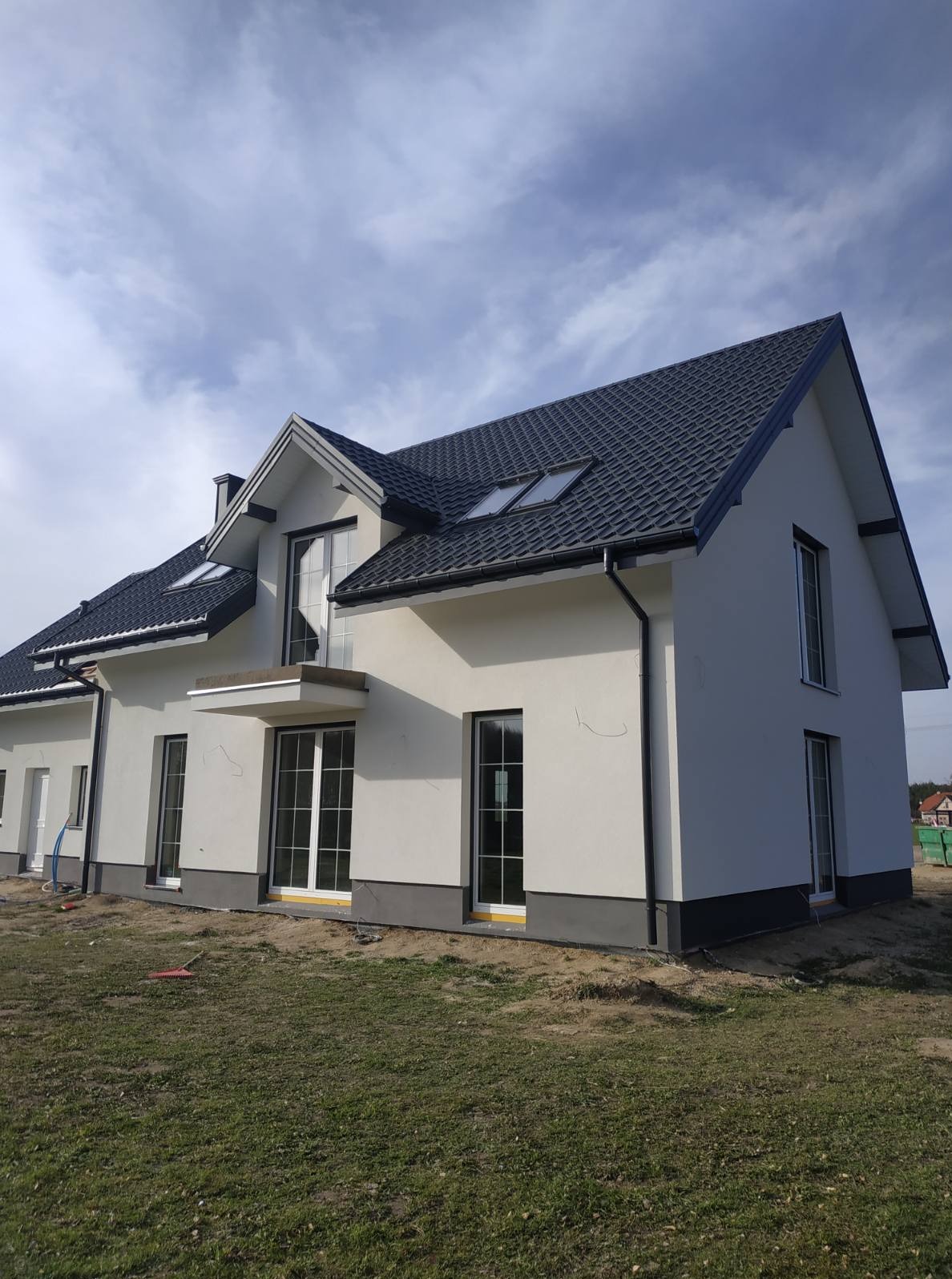 Nowoczesny dom jednorodzinny z jasną elewacją i ciemną dachówką, widok z boku, świeżo po tynkowaniu, z widocznymi detalami wykończeniowymi.