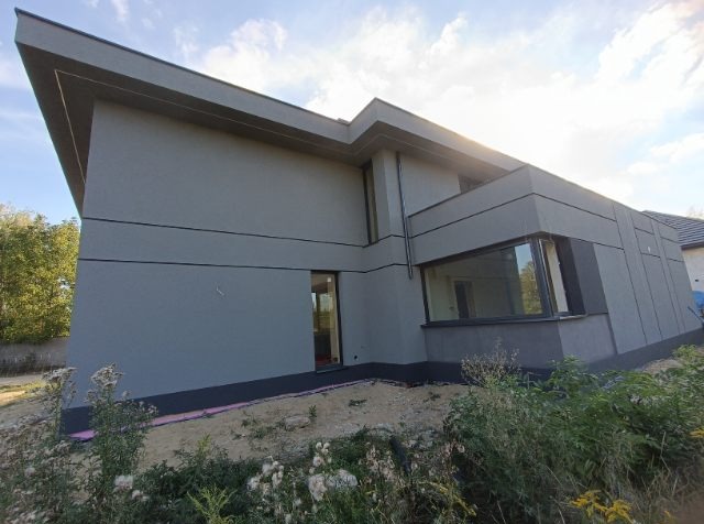 Nowoczesny dom z szarą elewacją, widoczne okna i fragment dachu. W tle zieleń, na pierwszym planie roślinność. Ujęcie z dołu.