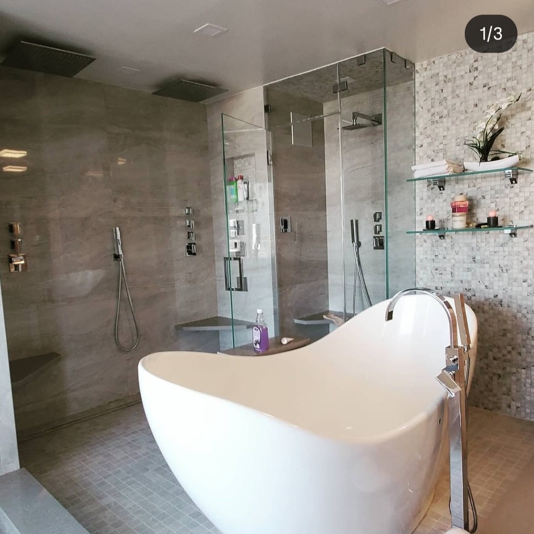 Nowoczesna łazienka z wolnostojącą wanną, szklanymi kabinami prysznicowymi i mozaiką na ścianie. Minimalistyczny design z elementami dekoracyjnymi na półkach.