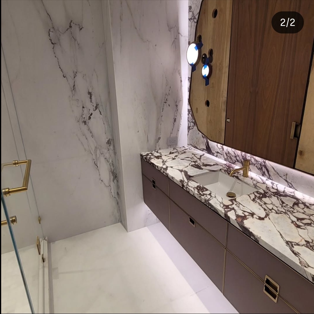 Elegancka łazienka z marmurowymi ścianami i blatem, brązowymi szafkami pod umywalką i złotą armaturą. Unikatowe oświetlenie i drewniane elementy dekoracyjne.