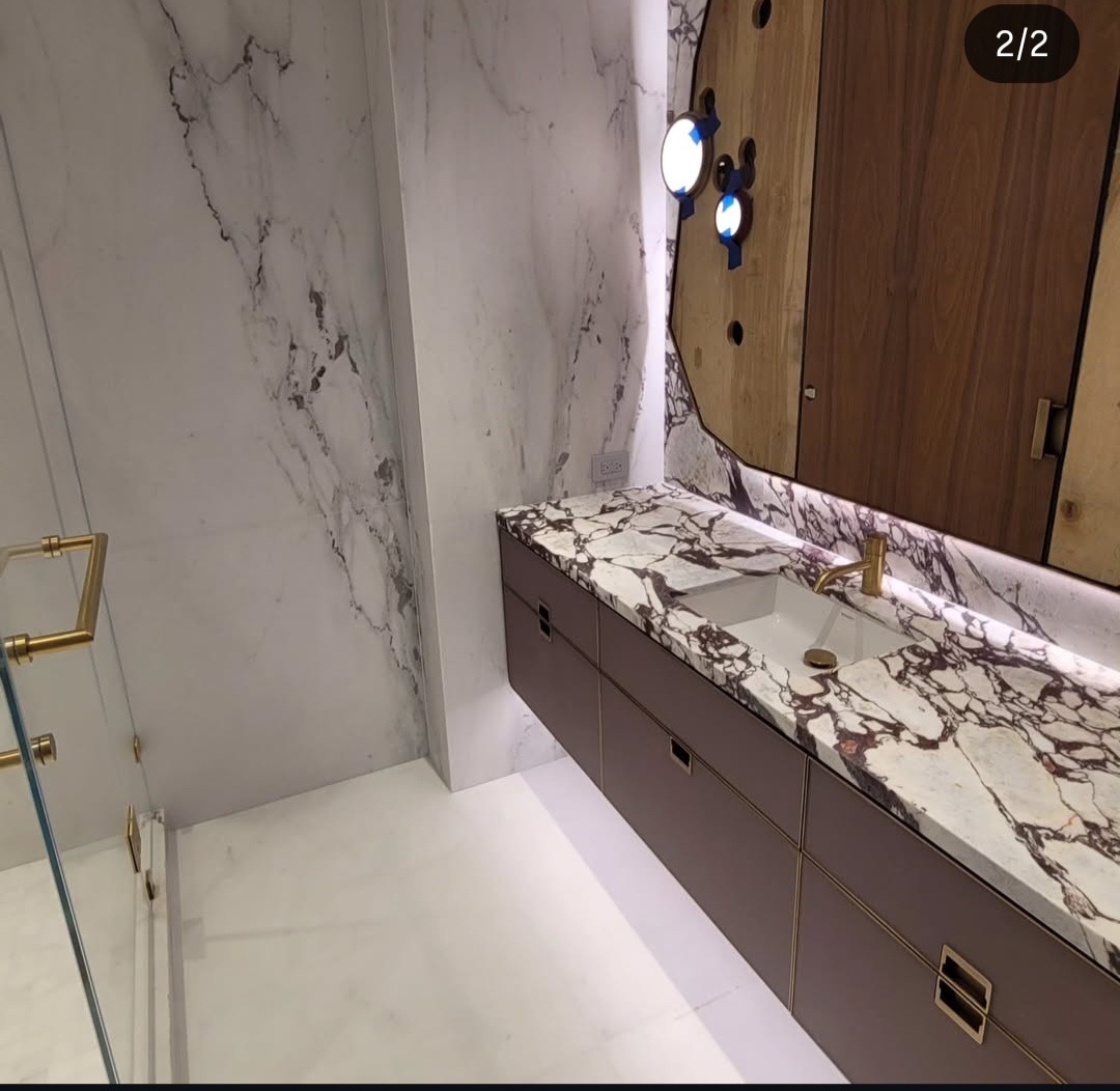 Elegancka łazienka z marmurową umywalką, złotą armaturą i drewnianą szafką z oświetleniem LED. Ściany wyłożone białym marmurem z szarymi żyłami.