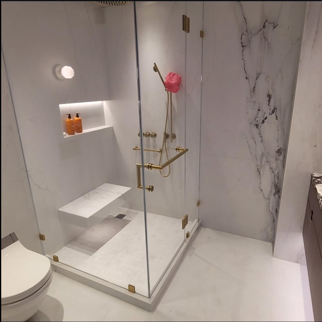 Nowoczesna kabina prysznicowa walk-in z siedziskiem, wykończona białym marmurem i złotymi detalami. Widoczne armatura, półka z kosmetykami i fragment toalety.