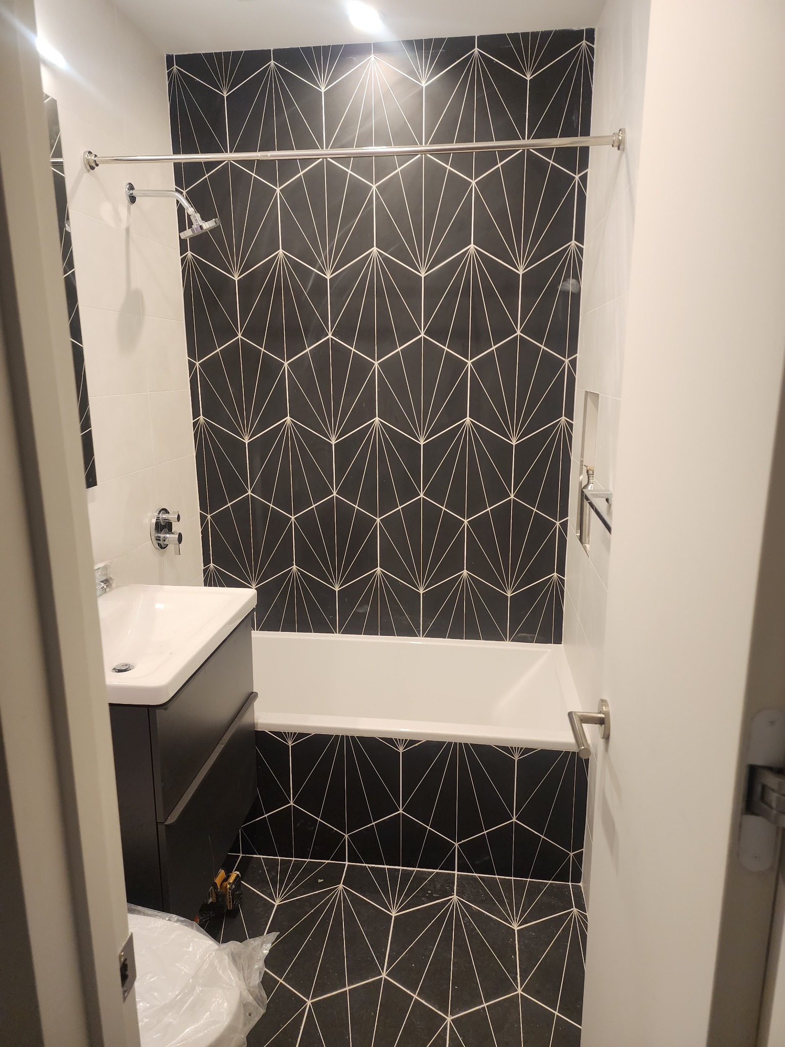 Wykończona łazienka z czarno-białymi geometrycznymi płytkami, białą wanną i umywalką. Widoczne narzędzia glazurnicze na podłodze. Nowoczesny design.