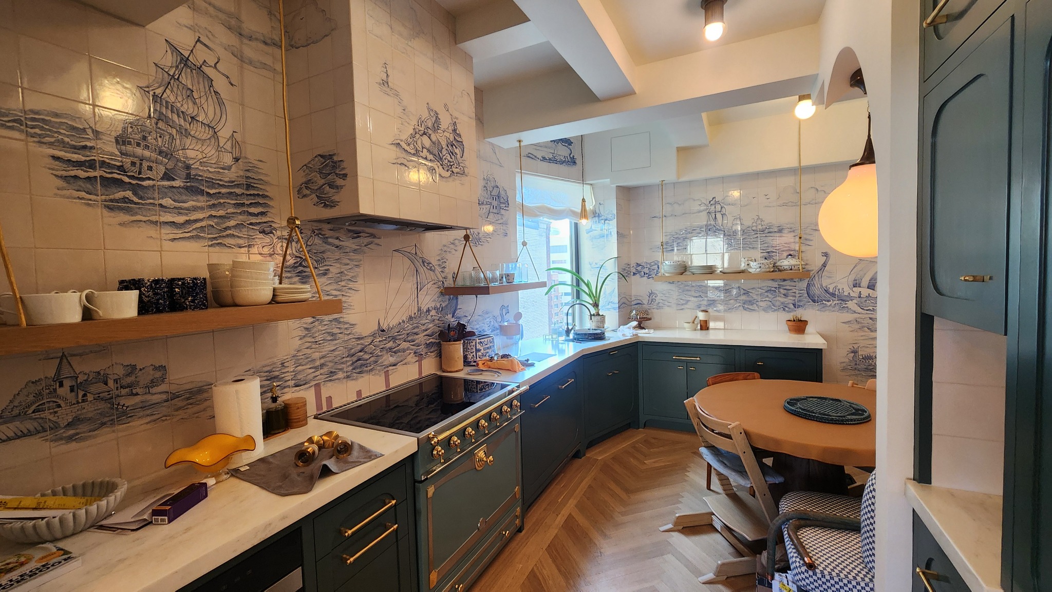 Wykończona kuchnia z płytkami w stylu holenderskim z motywem morskim, zielonymi szafkami i drewnianą podłogą w jodełkę. Widoczny fragment stołu i krzesła.