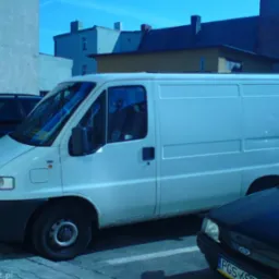Biały samochód dostawczy Fiat Ducato zaparkowany na parkingu miejskim, widoczny fragment czarnego samochodu osobowego z przodu.