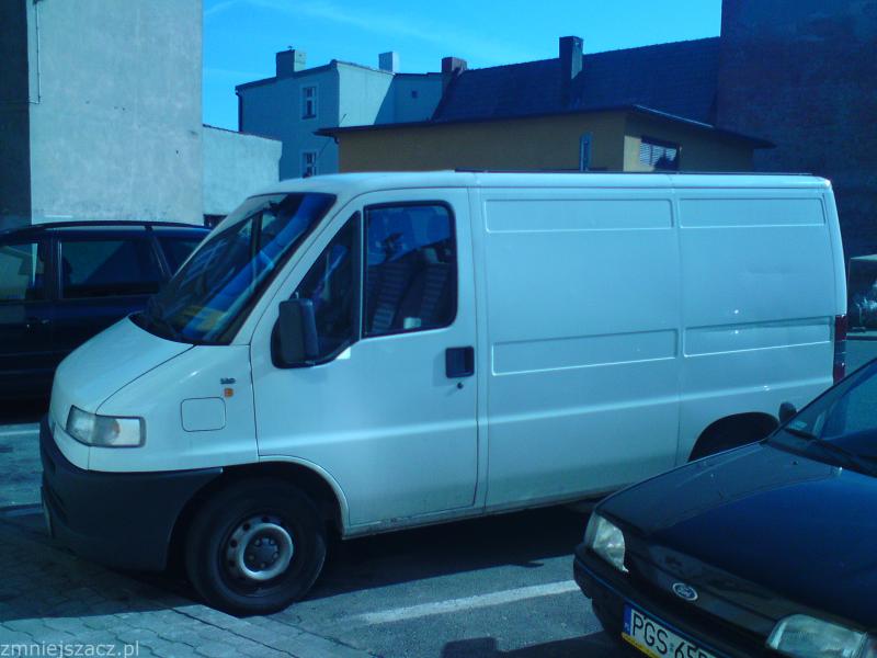 Biały samochód dostawczy Fiat Ducato zaparkowany na parkingu miejskim, widoczny fragment czarnego samochodu osobowego z przodu.