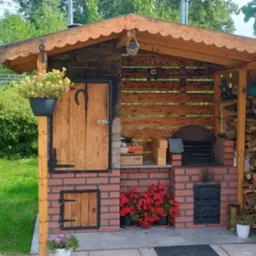 Zadaszona kuchnia letnia z grillem, z drewnianą konstrukcją i ceglanym murem, zdobiona kwiatami w doniczkach. W tle zieleń ogrodu.