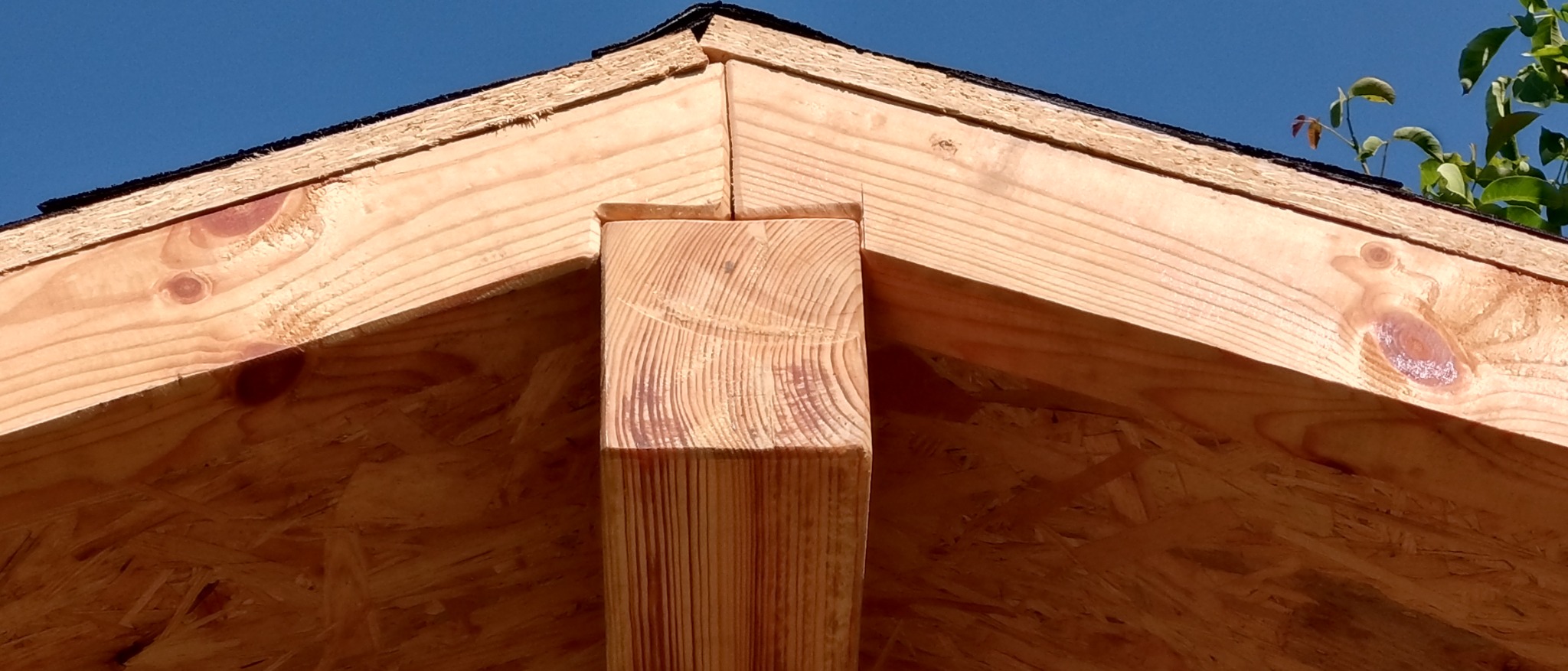 Fragment drewnianej konstrukcji dachu z widocznym słupem nośnym, deski i płyty OSB. Element konstrukcyjny na tle błękitnego nieba.