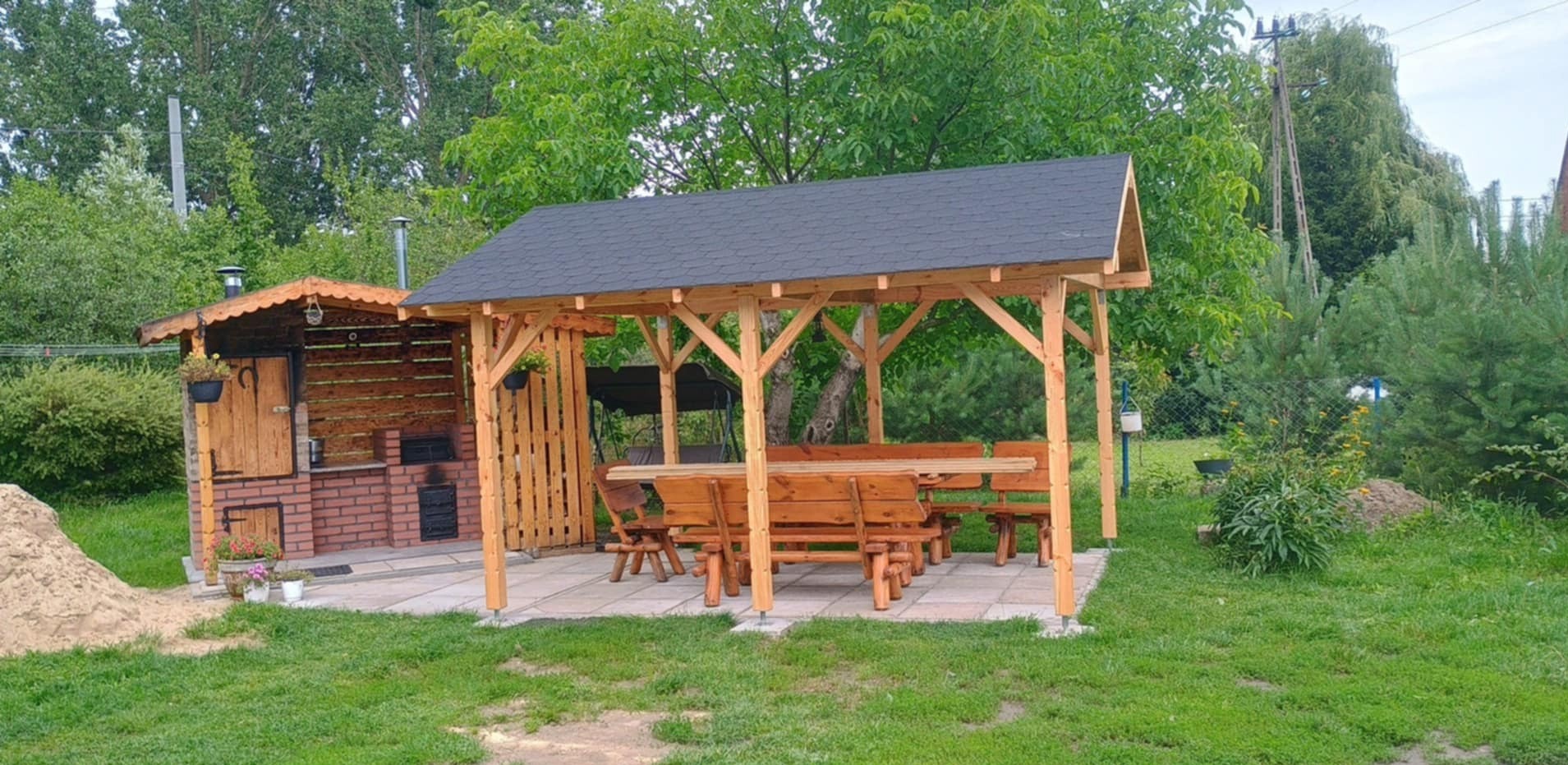 Drewniana pergola zadaszona gontem bitumicznym, z kompletem mebli ogrodowych i grillem z cegły na kamiennej posadzce. W tle zieleń ogrodu.