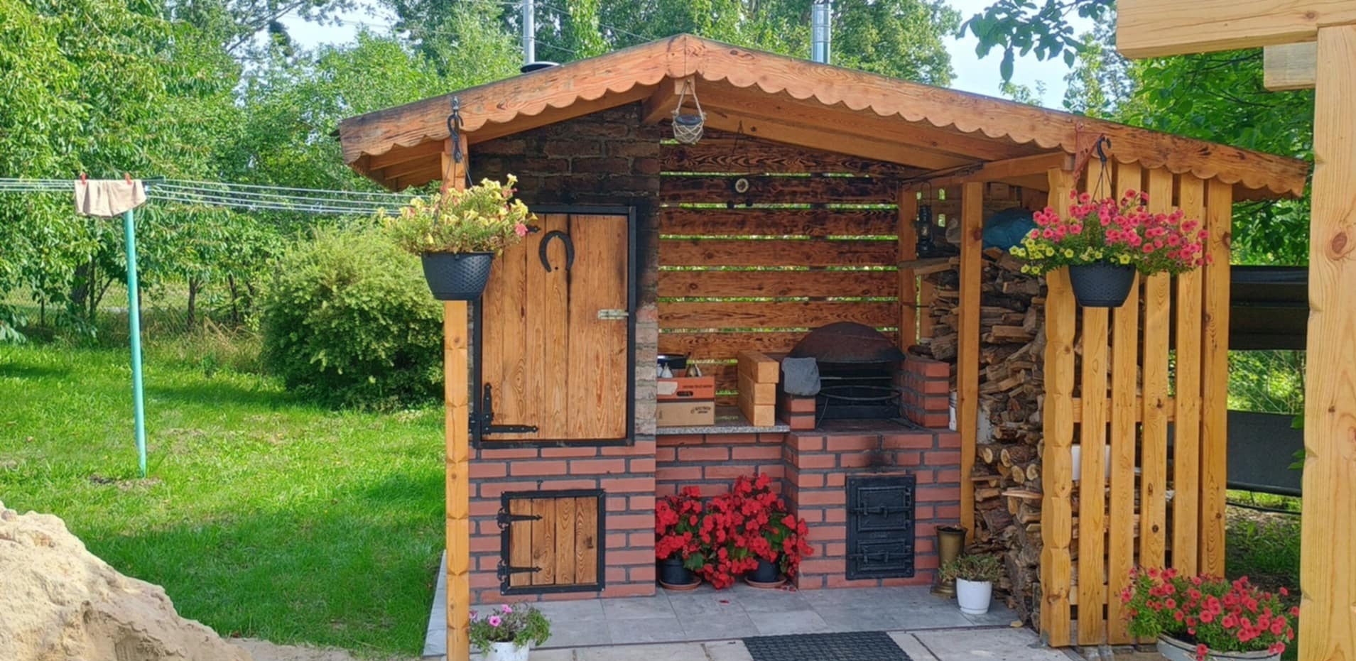 Zadaszona kuchnia letnia z grillem, z drewnianą konstrukcją i ceglanym murem, zdobiona kwiatami w doniczkach. W tle zieleń ogrodu.