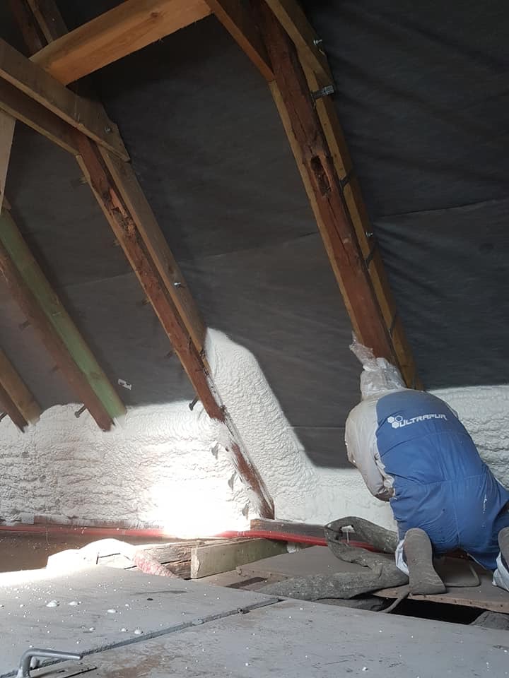 Pracownik w kombinezonie aplikuje pianę PUR na poddasze z drewnianą konstrukcją. Widoczna warstwa izolacji i elementy dachu. Oświetlenie punktowe.