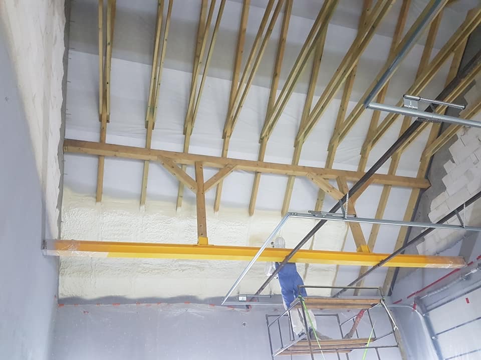 Poddasze z drewnianą konstrukcją dachu i ociepleniem pianką PUR; pracownik na rusztowaniu w trakcie aplikacji izolacji.