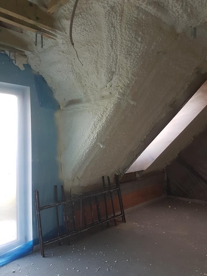 Poddasze po ociepleniu pianką PUR: biała izolacja pokrywa skośne ściany i sufit, widoczne okno dachowe i fragment konstrukcji więźby dachowej.