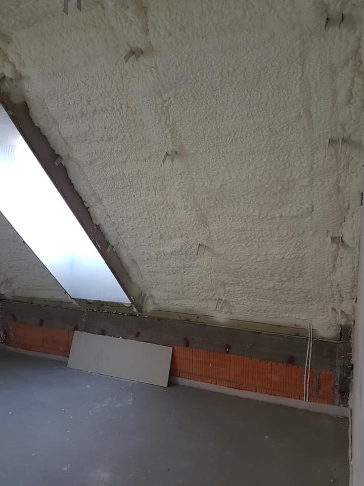 Poddasze po ociepleniu pianką PUR, widoczne okno dachowe i fragment ściany z cegły. Izolacja równomiernie rozprowadzona, szara podłoga.