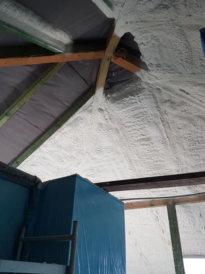 Poddasze po ociepleniu pianką PUR; widoczne drewniane elementy konstrukcyjne i charakterystyczna biała struktura pianki, fragment okna i zabezpieczony folią element.