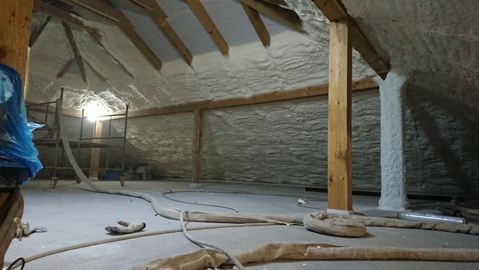 Poddasze po ociepleniu pianką PUR, widoczne drewniane elementy konstrukcyjne i szare węże na podłodze. Ściany i dach pokryte grubą warstwą izolacji.