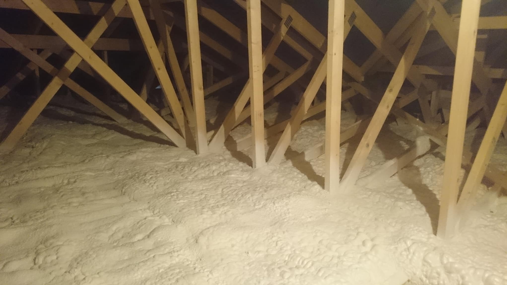 Poddasze z drewnianą konstrukcją wypełnione białą pianką izolacyjną, widok z góry, ciemne tło, oświetlenie punktowe.