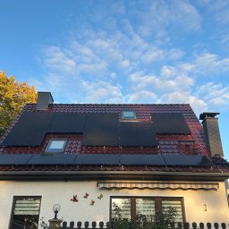 GROUP SLK SPÓŁKA Z OGRANICZONĄ ODPOWIEDZIALNOŚCIĄ - Czerwony dach z panelami fotowoltaicznymi w słoneczny dzień. Widoczne okna dachowe i dekoracje na elewacji domu. Jesienne drzewa w tle.