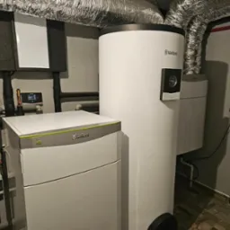 Biały zasobnik ciepłej wody Vaillant i pompa ciepła Green iQ w kotłowni z rurami i klimatyzacją. Widok z boku na urządzenia.