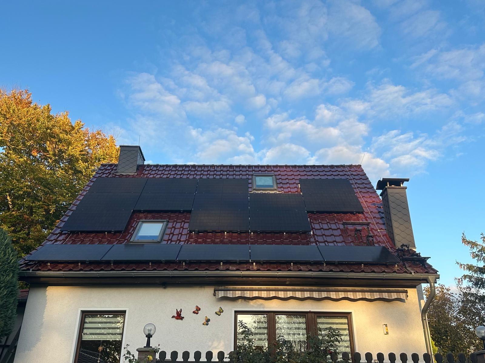 Czerwony dach z panelami fotowoltaicznymi w słoneczny dzień. Widoczne okna dachowe i dekoracje na elewacji domu. Jesienne drzewa w tle.