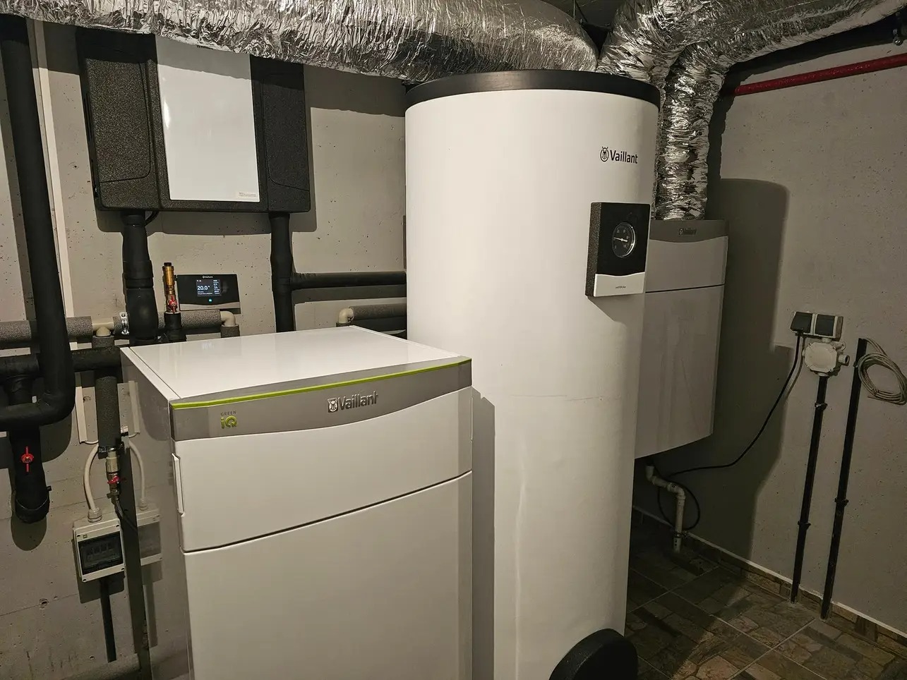 Biały zasobnik ciepłej wody Vaillant i pompa ciepła Green iQ w kotłowni z rurami i klimatyzacją. Widok z boku na urządzenia.