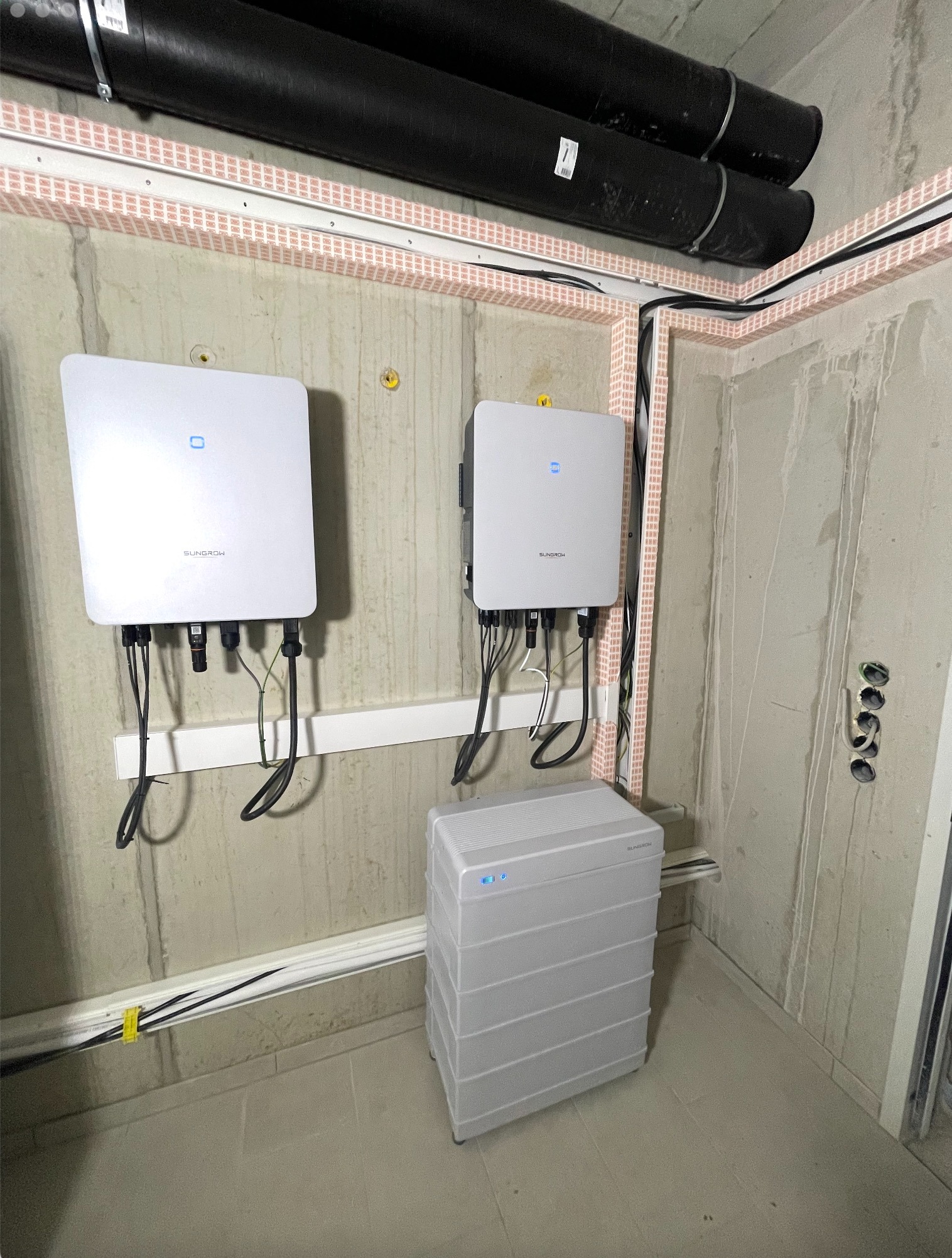 Dwa falowniki i magazyn energii Sungrow zamontowane na surowej ścianie. Widoczne rury instalacyjne pod sufitem i okablowanie. Detal: żółte oznaczenia na kablach.