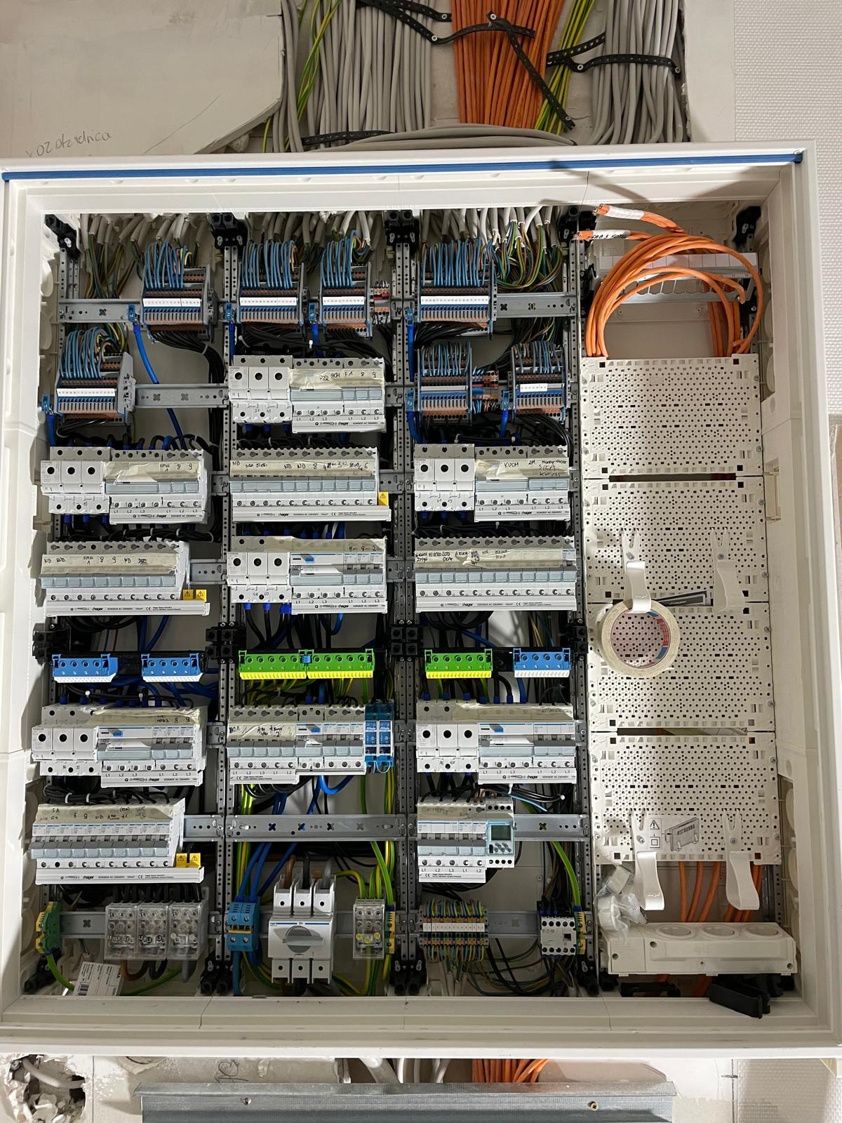 Rozdzielnica elektryczna w zabudowie, widoczne liczne kable, bezpieczniki i elementy montażowe. Pomarańczowe kable ułożone po prawej stronie. Widok z bliska.