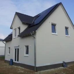 Nowoczesny dom z jasną elewacją i panelami słonecznymi na dachu, widoczny z perspektywy narożnej. W tle błękitne niebo, obok domu zaparkowany bus.