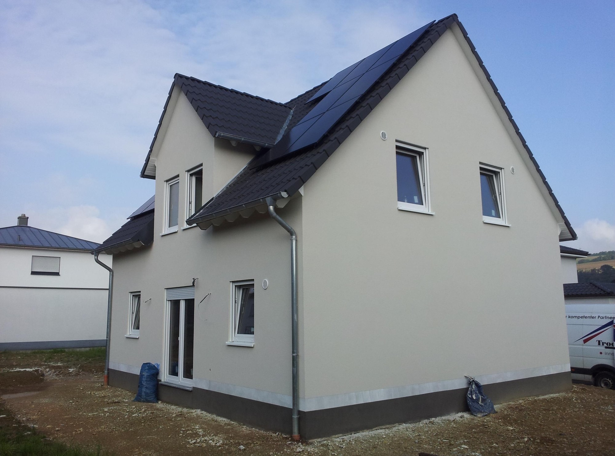 Nowoczesny dom z jasną elewacją i panelami słonecznymi na dachu, widoczny z perspektywy narożnej. W tle błękitne niebo, obok domu zaparkowany bus.