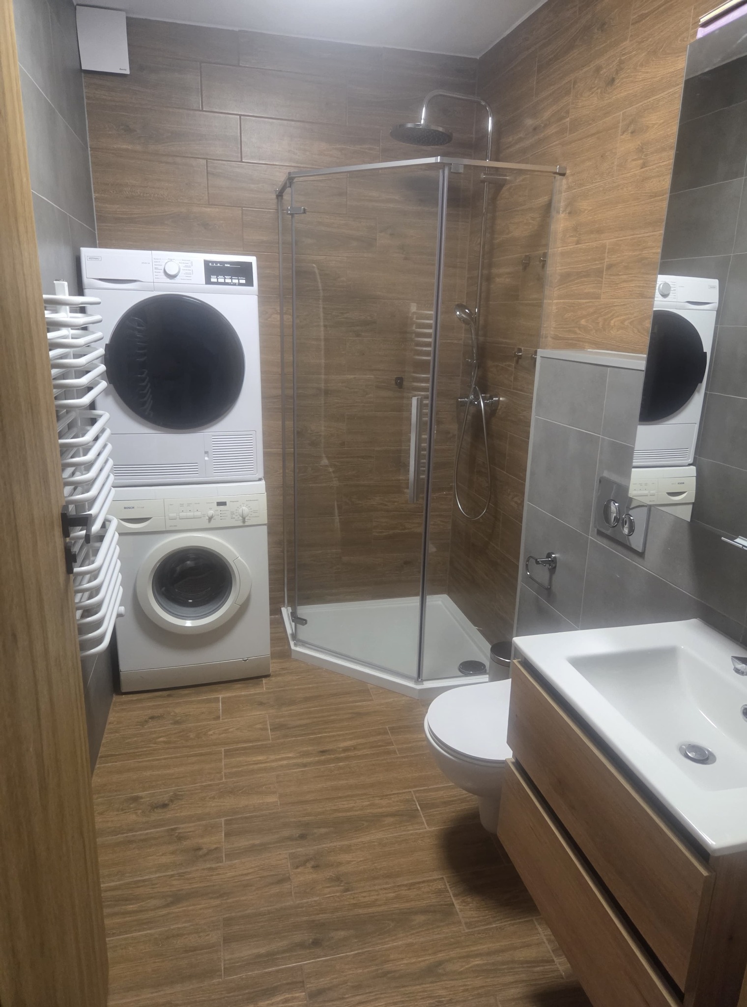 Nowoczesna łazienka z kabiną prysznicową, umywalką z szafką i pralko-suszarką w zabudowie. Płytki imitujące drewno na podłodze i ścianach. Minimalistyczny design.