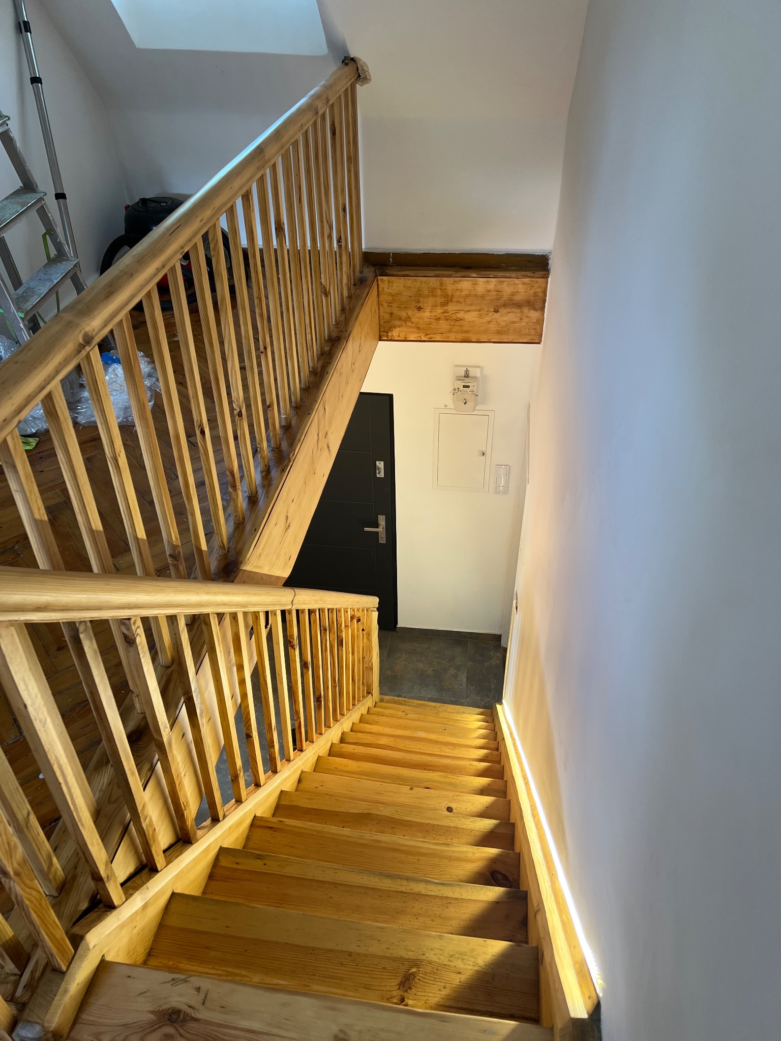 Drewniane schody z balustradą po renowacji, widok z góry. Jasne drewno, białe ściany, czarne drzwi w tle. Schody prowadzą w dół, oświetlone naturalnym światłem.
