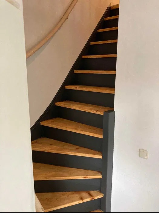 Odnowione drewniane schody z czarną balustradą i naturalnym drewnem stopni, oświetlone naturalnym światłem. Widoczny fragment drewnianej poręczy.