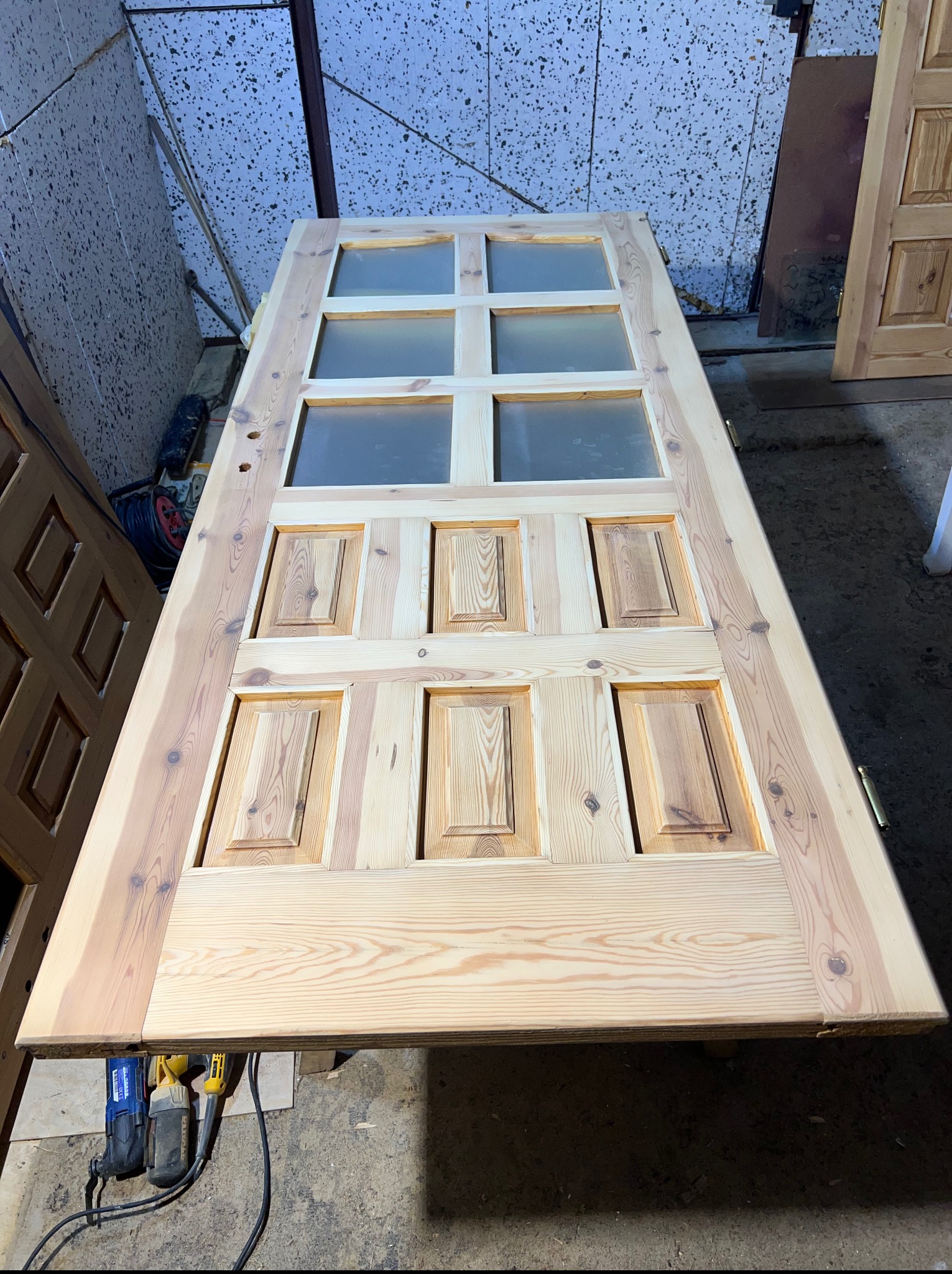 Odrestaurowane drewniane drzwi z szybami, leżące poziomo na stole warsztatowym, z widocznymi narzędziami szlifierskimi i elementami wykończenia. Jasne drewno po renowacji.