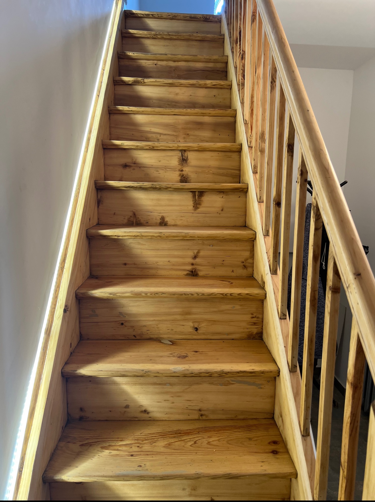 Naturalne drewniane schody z balustradą, oświetlone od góry, ukazane z dołu. Widoczne słoje drewna i detale konstrukcyjne.