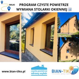 BIAN-TIKO MACIEJ KIERZNOWSKI - Żółty dom po wymianie okien w ramach programu Czyste Powietrze. Widoczne nowe okna z brązowymi ramami i odnowiona elewacja. Logo Bian-Tik i adres strony internetowej.