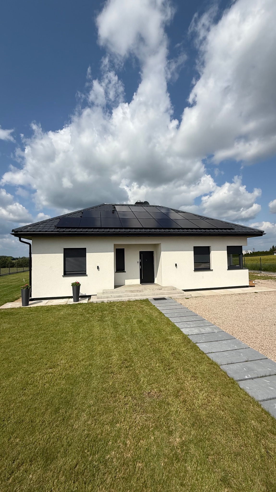 Biały dom z panelami słonecznymi na dachu, widok z przodu, trawnik i ścieżka z płyt. Błękitne niebo z obłokami w tle. Nowoczesna architektura.