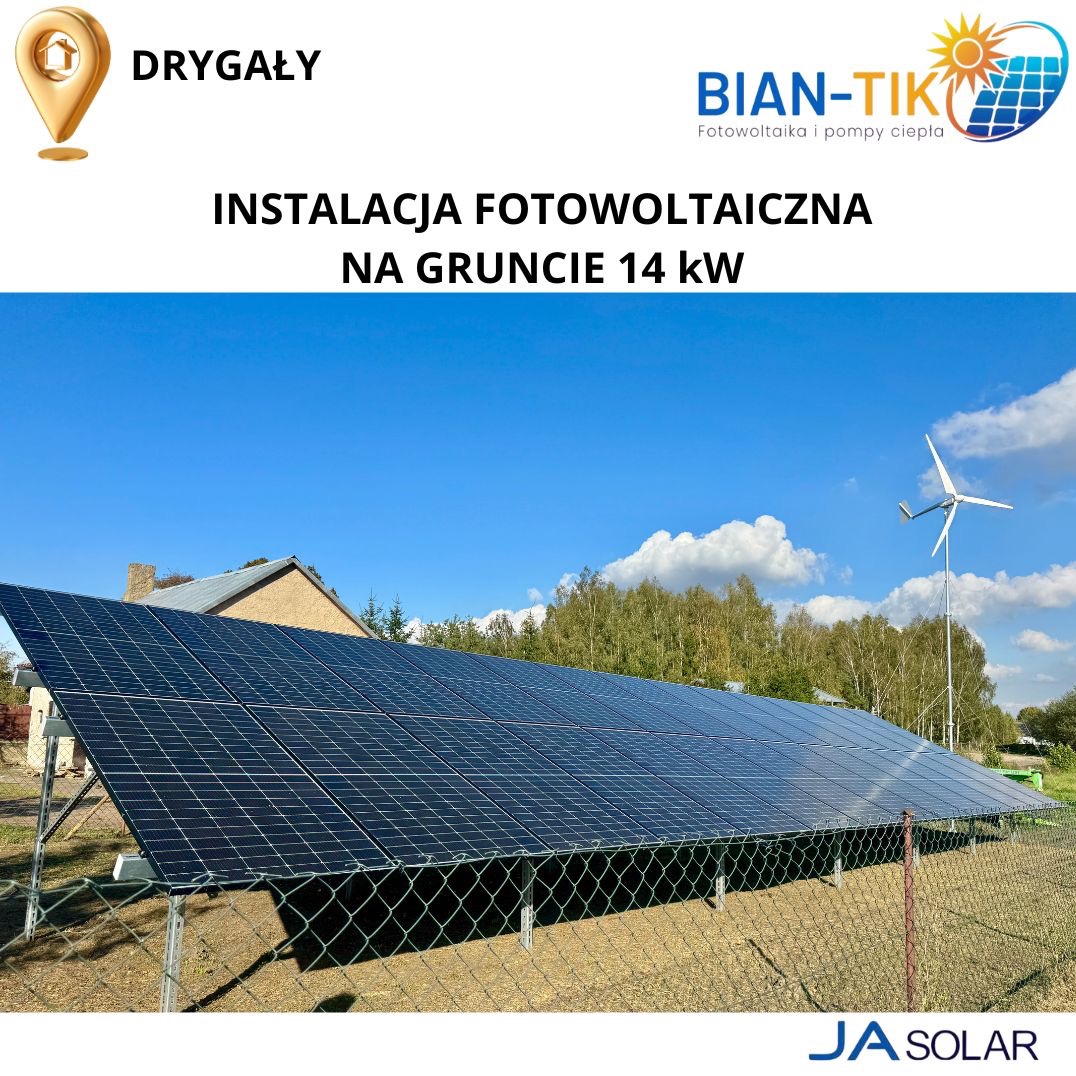 Instalacja fotowoltaiczna na gruncie o mocy 14 kW, widoczna zza ogrodzenia, z turbiną wiatrową w tle pod błękitnym niebem.