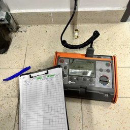 Usługi Elektryczne i Teletechniczne - Miernik elektryczny Sonel MPL-520 podłączony do gniazdka, obok notatnik z zapisanymi wynikami pomiarów. Widok z góry na kafelkową podłogę.