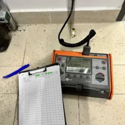 Miernik elektryczny Sonel MPL-520 podłączony do gniazdka, obok notatnik z zapisanymi wynikami pomiarów. Widok z góry na kafelkową podłogę.