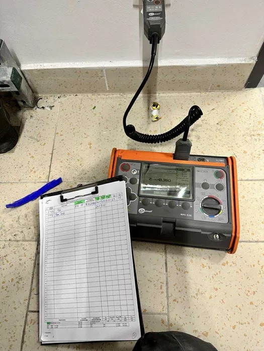 Miernik elektryczny Sonel MPL-520 podłączony do gniazdka, obok notatnik z zapisanymi wynikami pomiarów. Widok z góry na kafelkową podłogę.