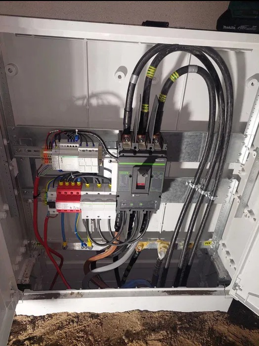 Otwarta skrzynka elektryczna z zainstalowanym wyłącznikiem, przewodami i zabezpieczeniami. Widoczne elementy instalacji elektrycznej wewnątrz obudowy.