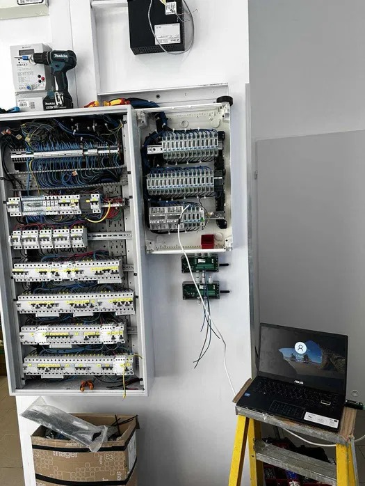 Rozdzielnica elektryczna w trakcie montażu, z widocznymi przewodami, zabezpieczeniami i laptopem na drabinie. Widoczny również miernik i wkrętarka.
