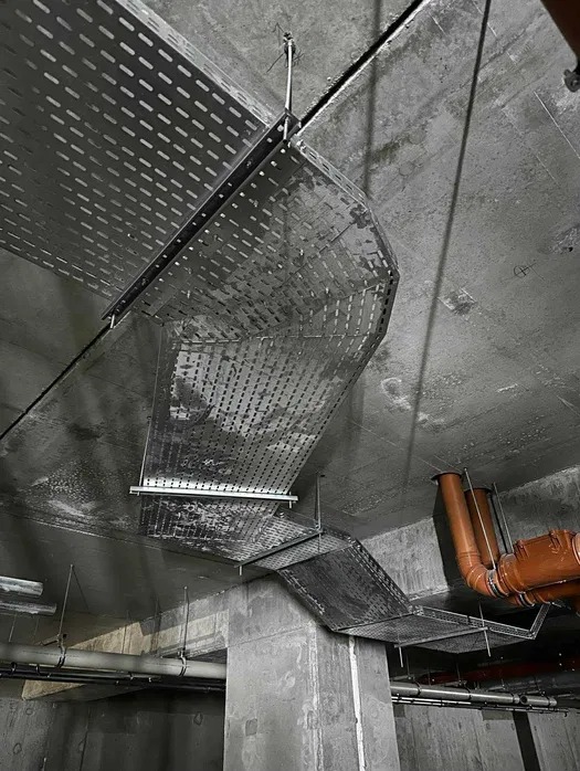 Metalowe korytka kablowe z perforacją, zamontowane pod betonowym sufitem. Widoczne rury instalacyjne w tle. Surowy, industrialny charakter wnętrza.