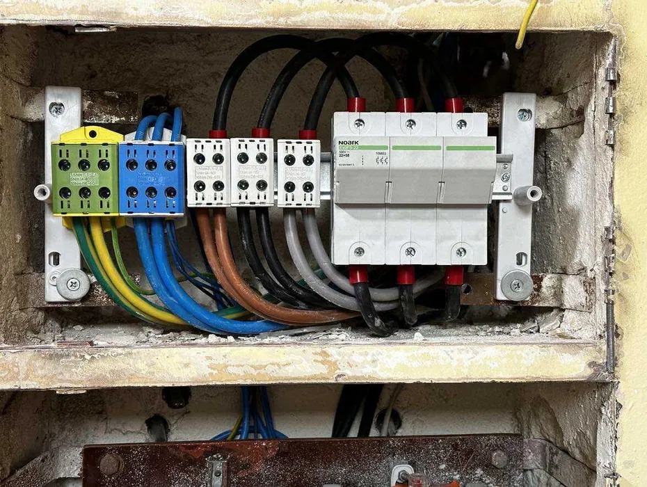 Rozdzielnica elektryczna z kolorowymi zaciskami (zielony, niebieski, biały) i zabezpieczeniami przepięciowymi NOARK, zamontowana w starej, betonowej wnęce.