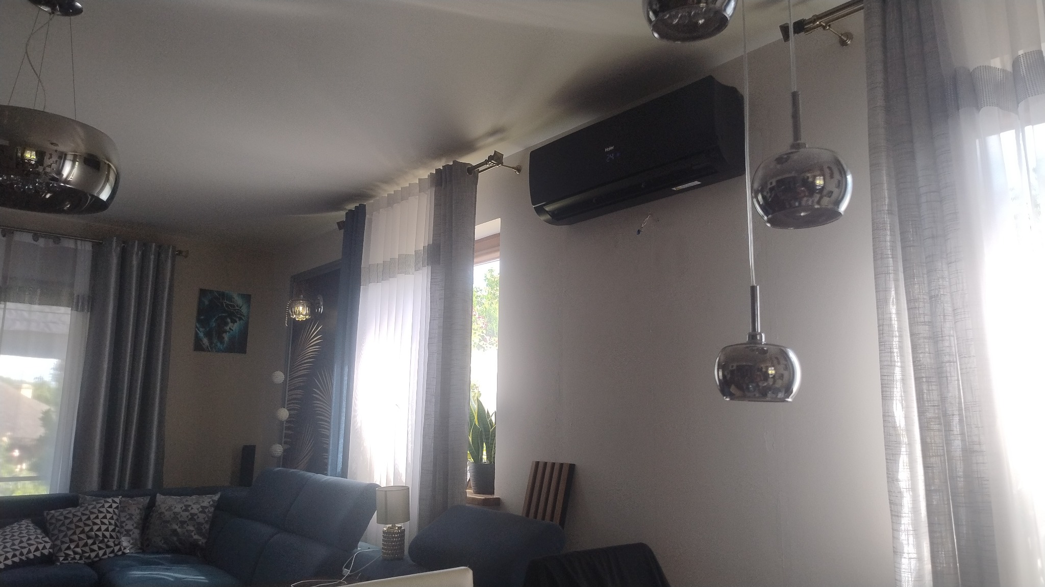 Czarny klimatyzator Haier zamontowany na ścianie w salonie. Widoczne srebrne lampy wiszące i zasłony w oknie. Nowoczesny design.