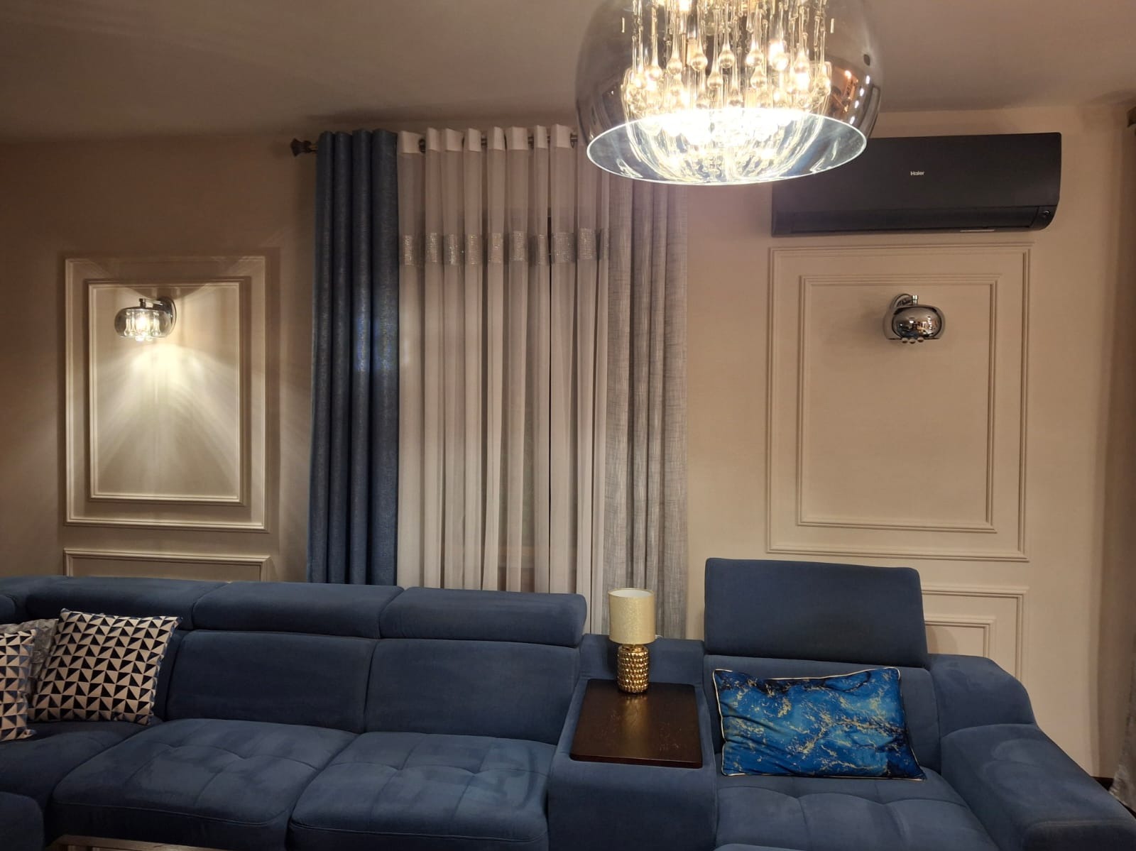 Nowoczesny salon z klimatyzacją Haier w kolorze czarnym, zamontowaną nad ozdobną listwą. Wnętrze dopełnia niebieska sofa i kryształowy żyrandol.