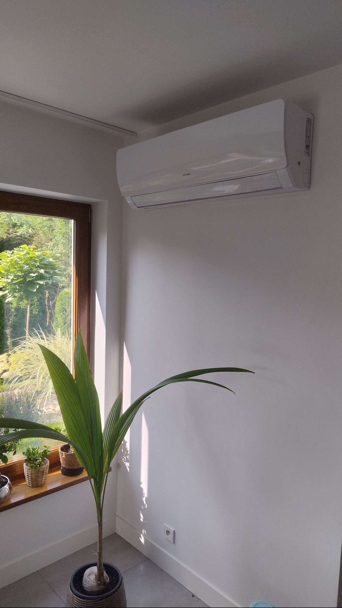 Biały klimatyzator ścienny w jasnym wnętrzu, widok z dołu, obok okno z zielenią i roślina doniczkowa. Czysty montaż, minimalistyczny design.